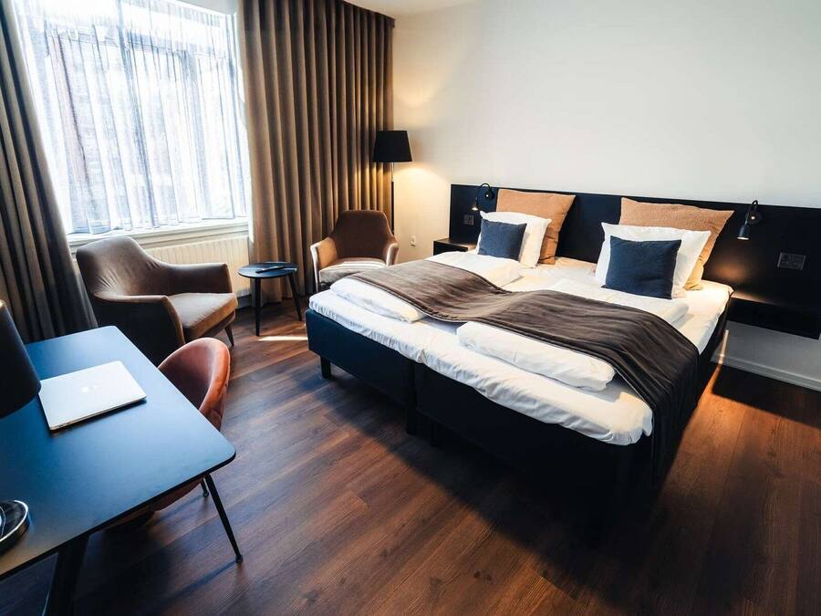 Habitación de hotel moderna con una cama grande, un escritorio con una computadora portátil, una silla y una ventana con cortinas.