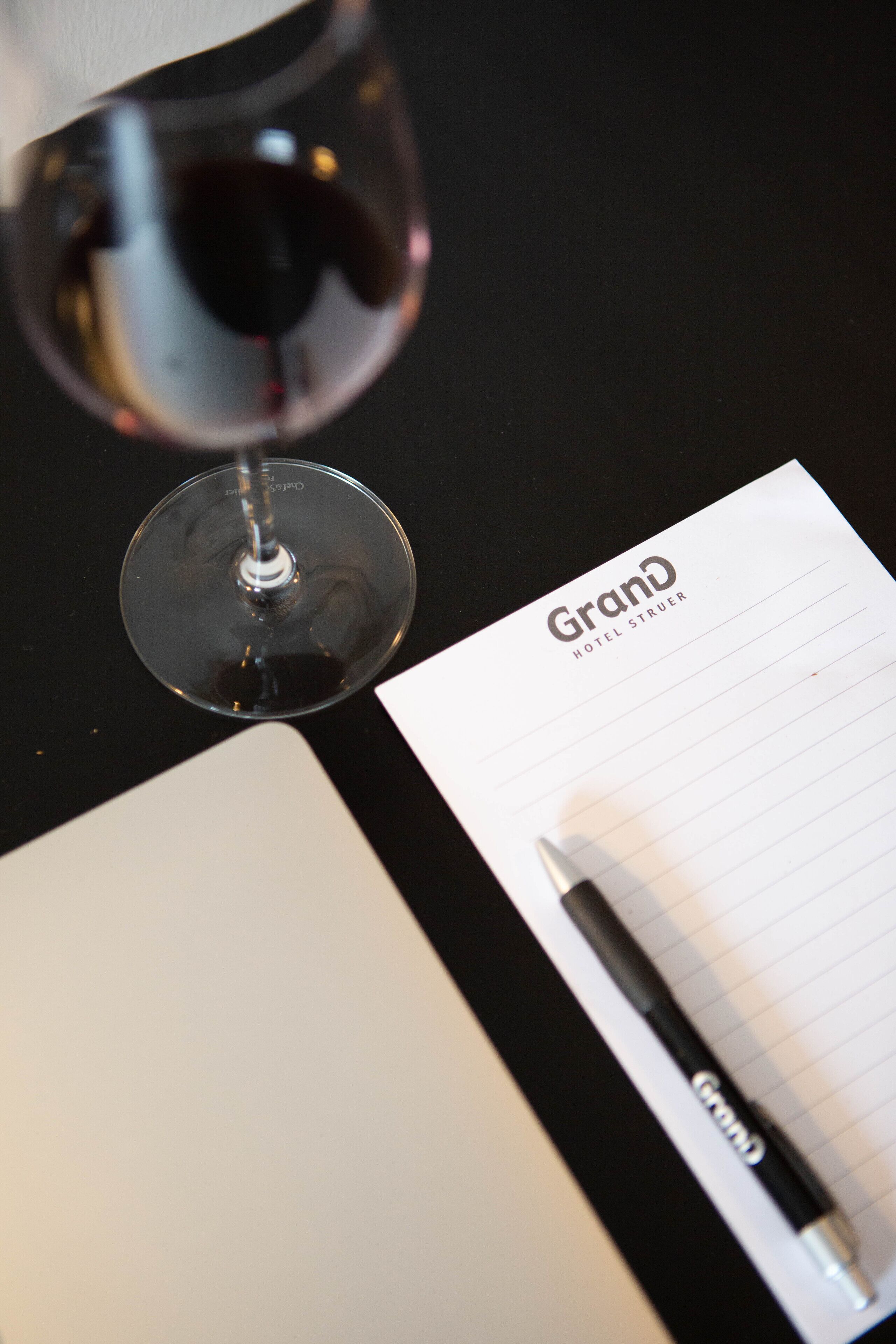 A wine glass, a notepad with the word "Grand" on it, and a pen on a black surface.