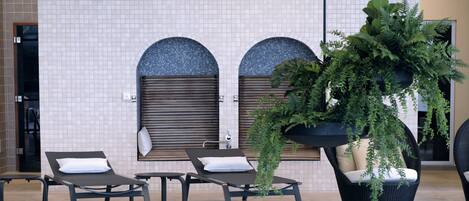 Un espace aménagé au bord de la piscine, avec des chaises longues, un mur recouvert de carreaux et agrémenté d’arcs, ainsi qu’une grande plante en pot.
