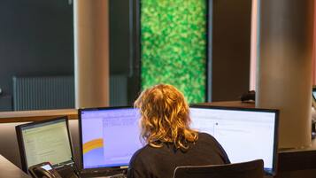 En person arbejder ved et skrivebord med flere computerskærme, en telefon og papirer.