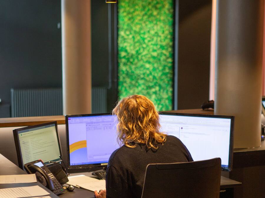 En person arbejder ved et skrivebord med flere computerskærme, en telefon og papirer.