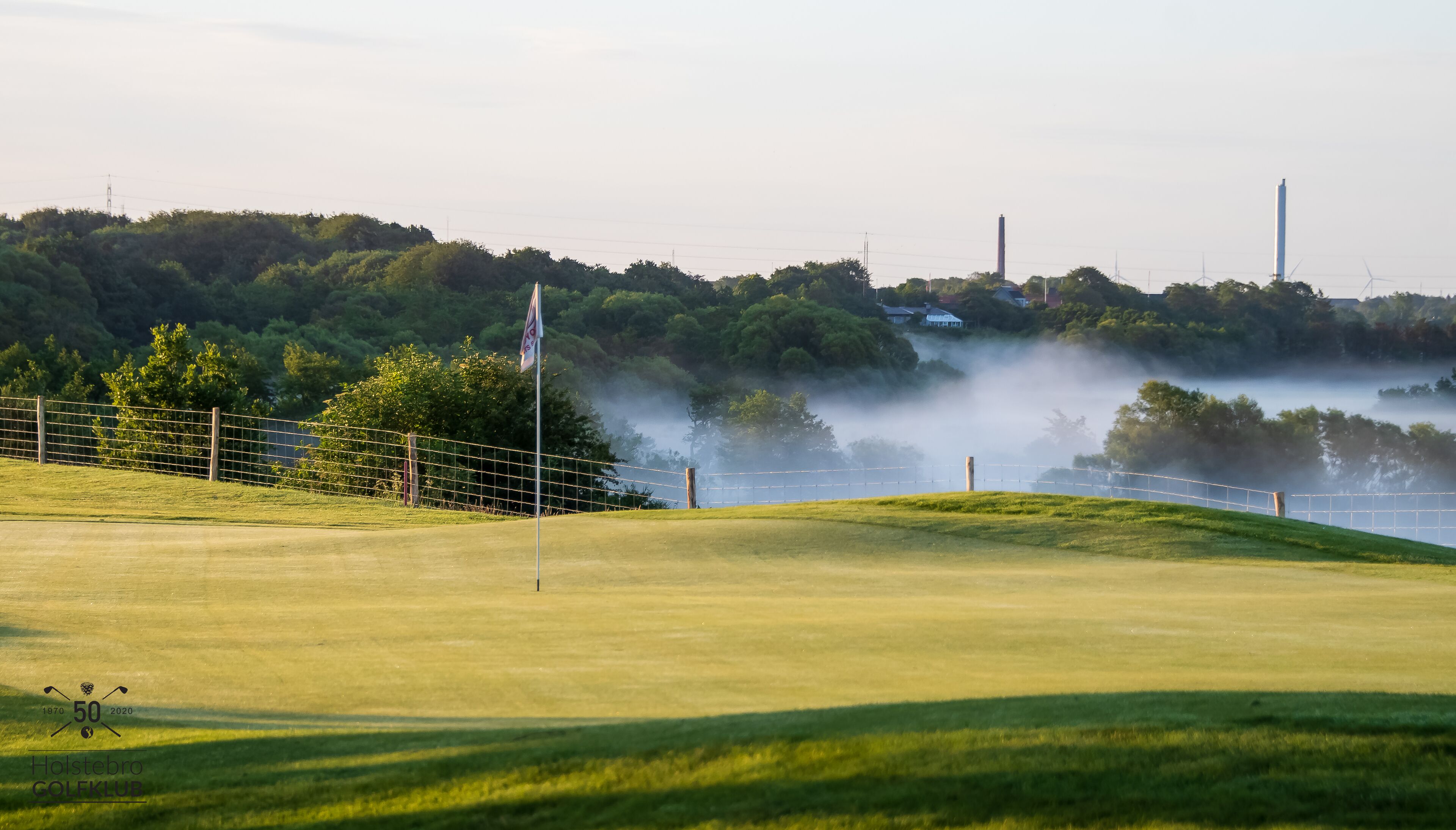 A golf course with a flag, a fence, and a misty background with trees and industrial structures.