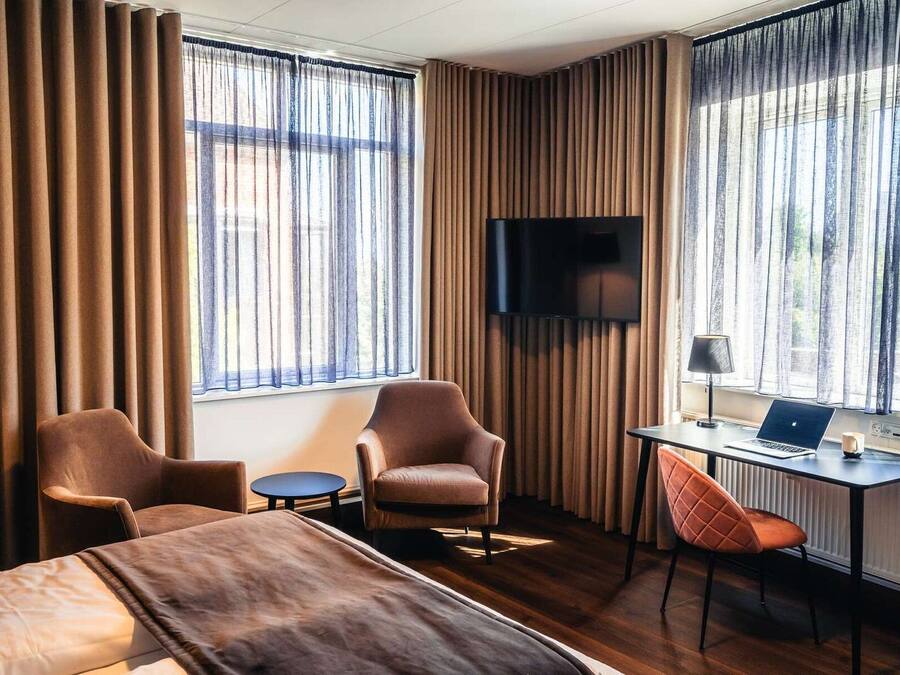 Et hotelværelse med en seng, to lænestole, et skrivebord med en bærbar computer og et fjernsyn ophængt på væggen.