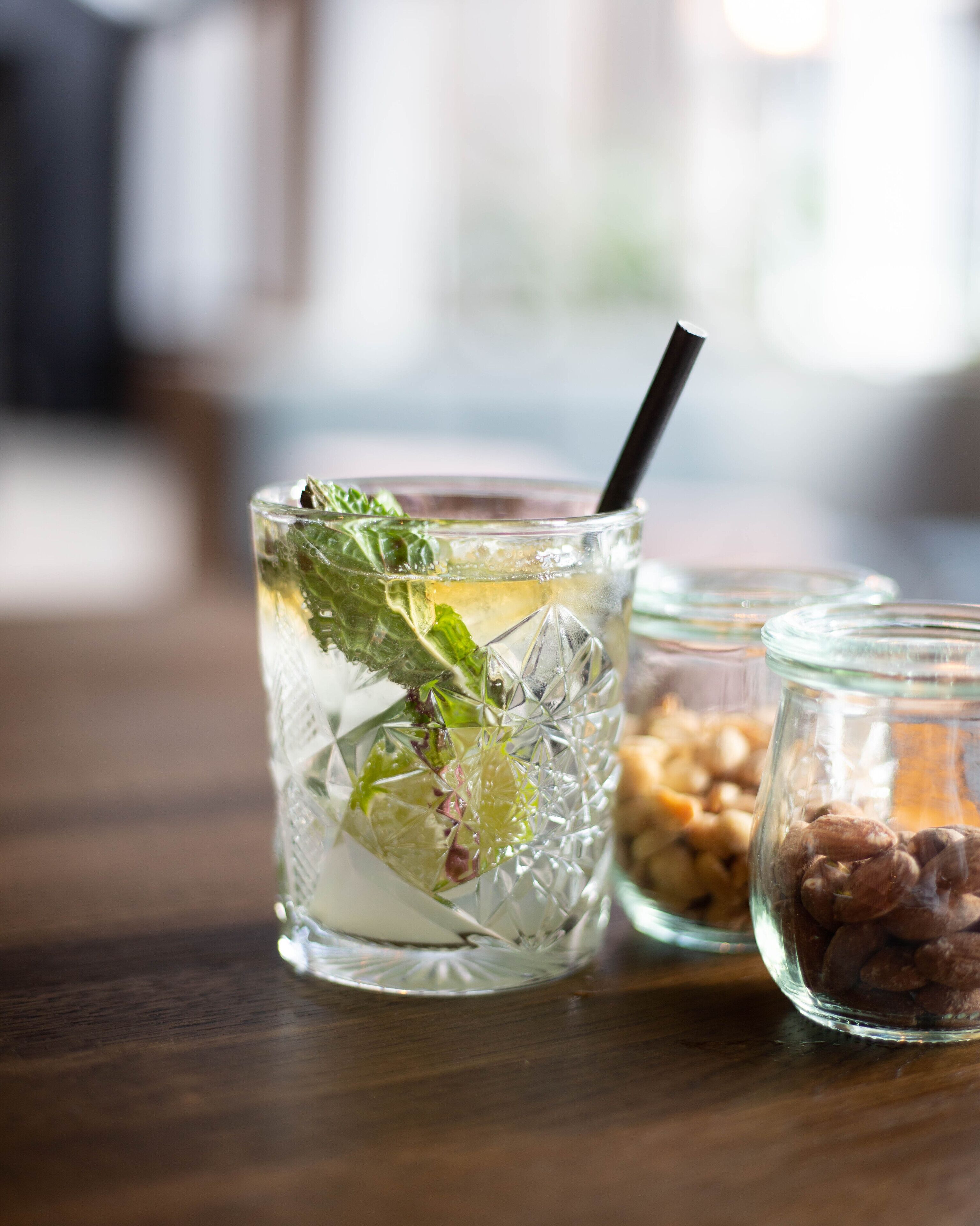 A glass of iced beverage with mint leaves, a black straw, and a jar of nuts on a wooden table.