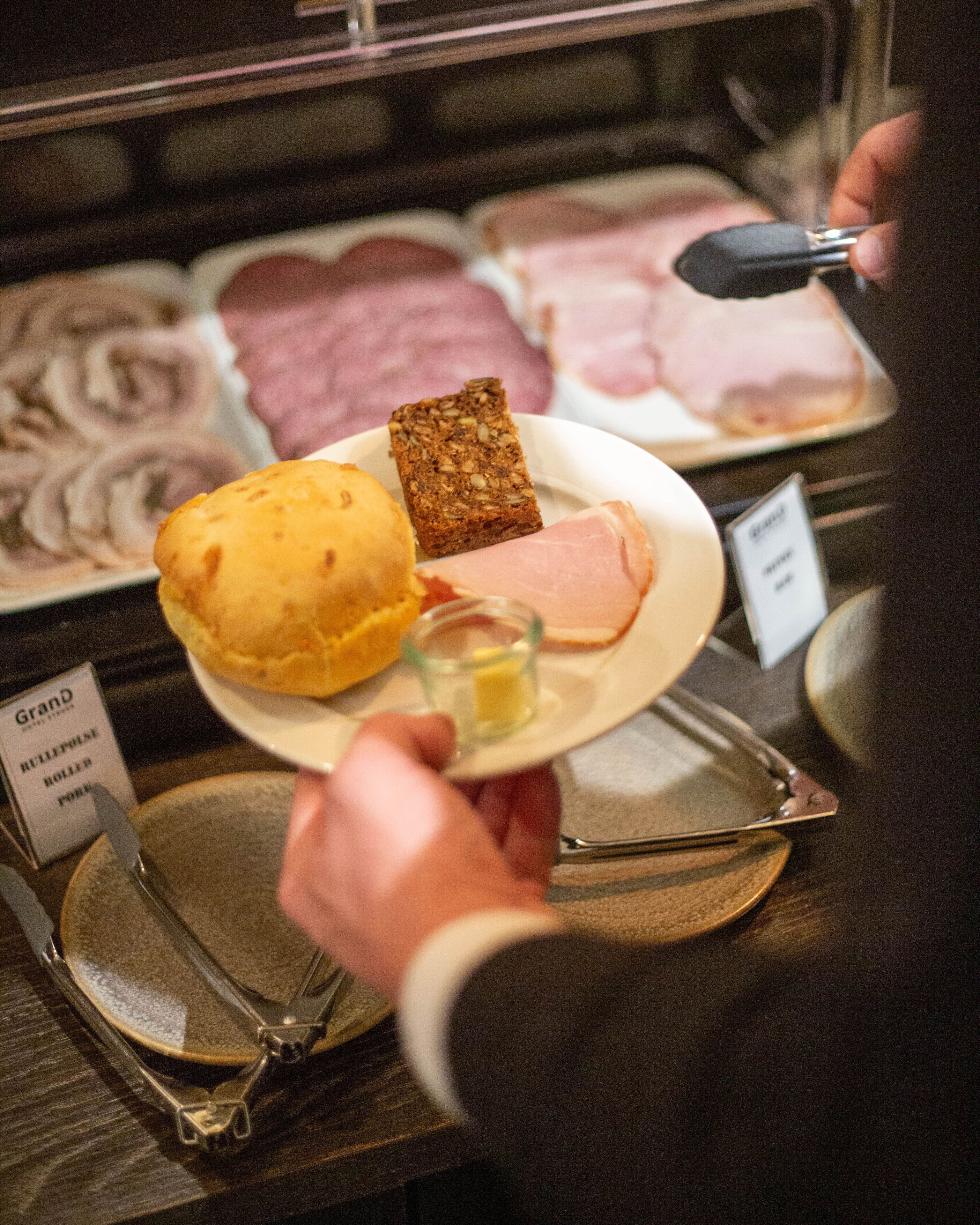 Una persona sosteniendo un plato con pan, jamón y un pequeño recipiente de vidrio, probablemente mantequilla, en un deli o panadería.