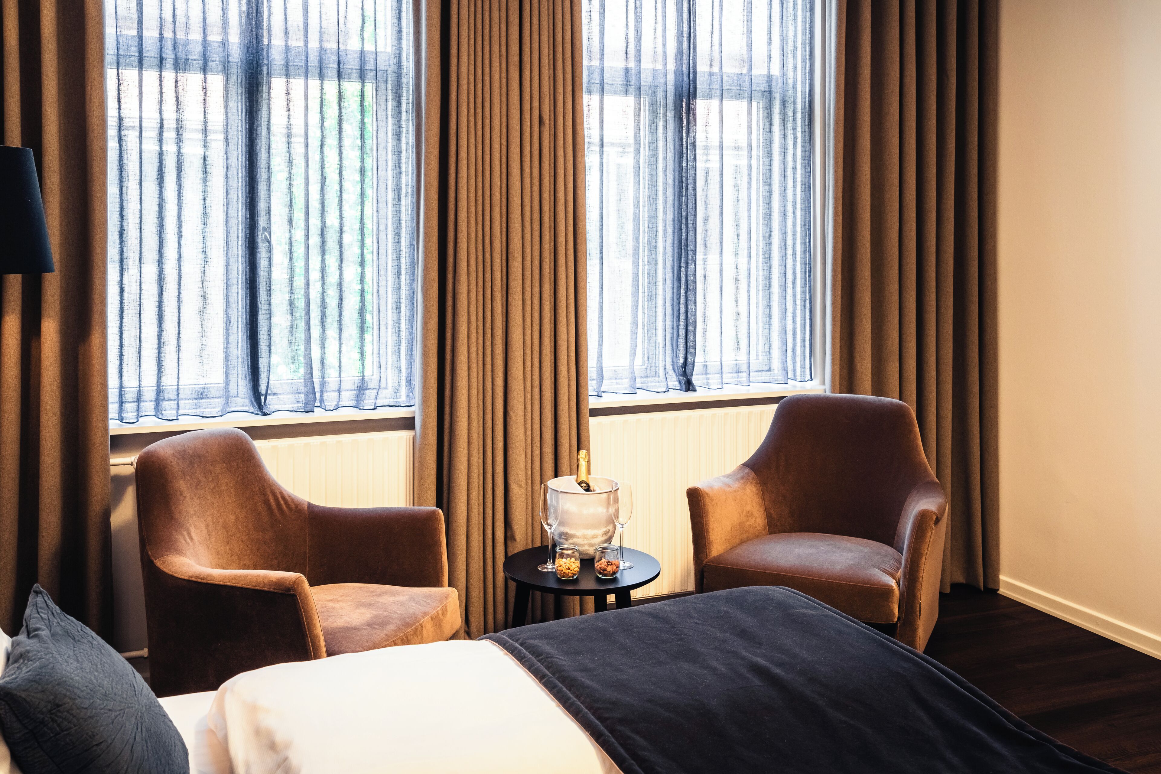A hotel room with a bed, two armchairs, a small table with a bottle and glasses, and a view through sheer curtains.