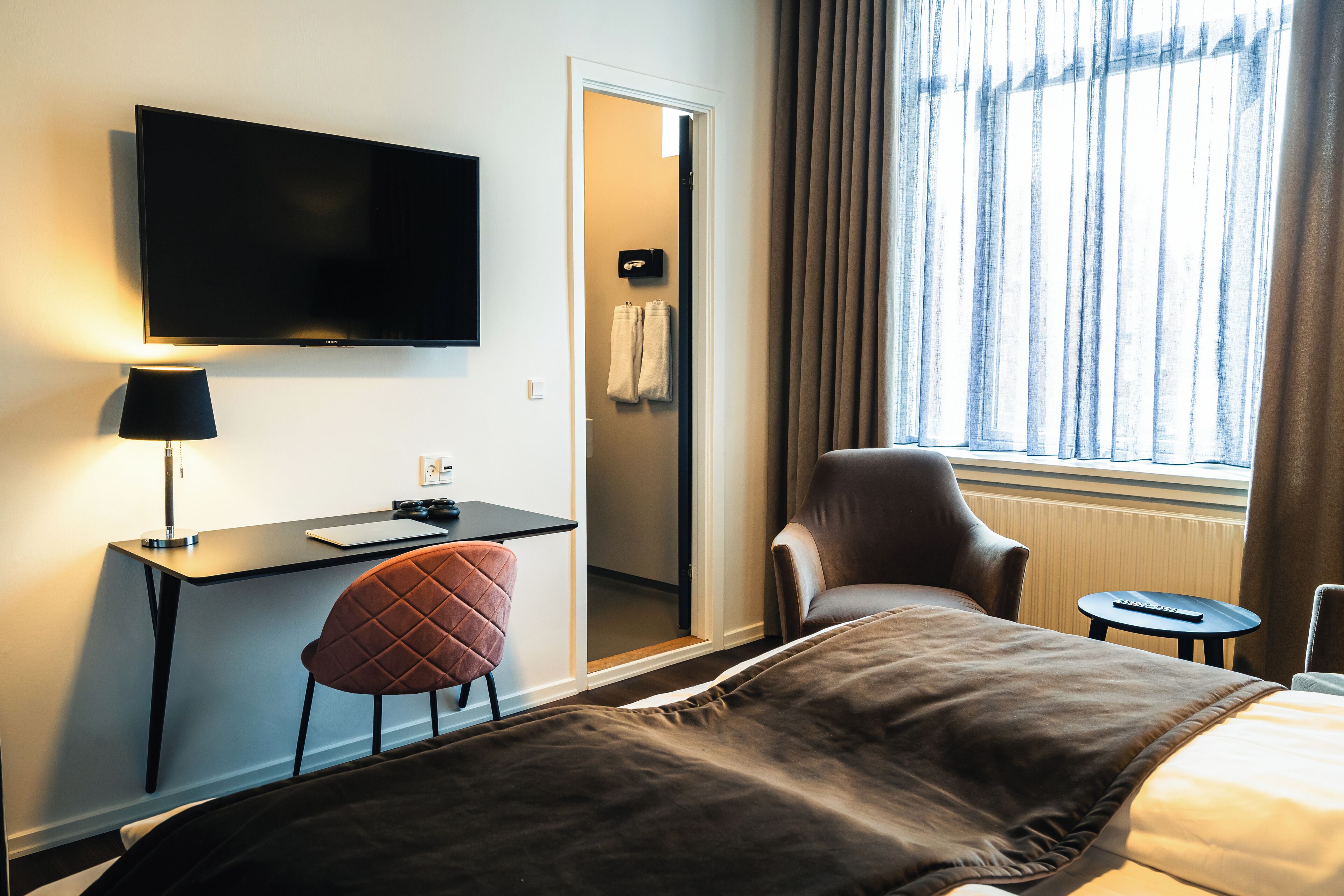 Habitación de hotel con cama, escritorio con computadora, televisor en la pared, una silla y ventana con cortinas.