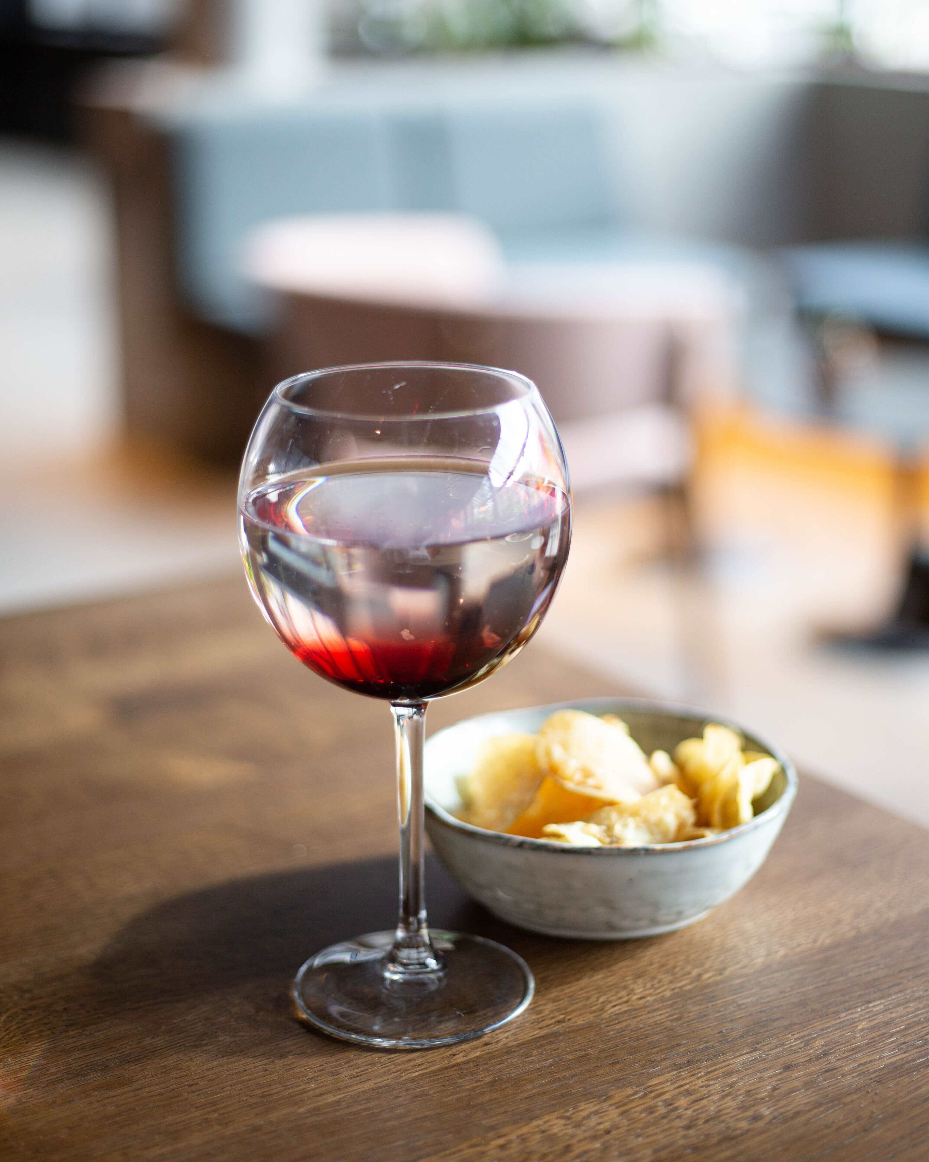 A glass of red wine on a wooden table with a bowl of snacks beside it.