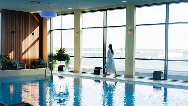 An indoor swimming pool with a view of the water outside, large windows, and a woman walking. - Grand Hotel Struer (Struer)