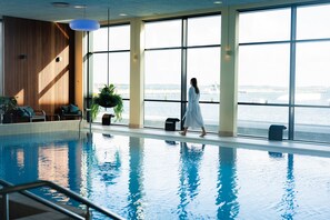 An indoor swimming pool with a view of the water outside, large windows, and a woman walking. - Grand Hotel Struer (Struer)