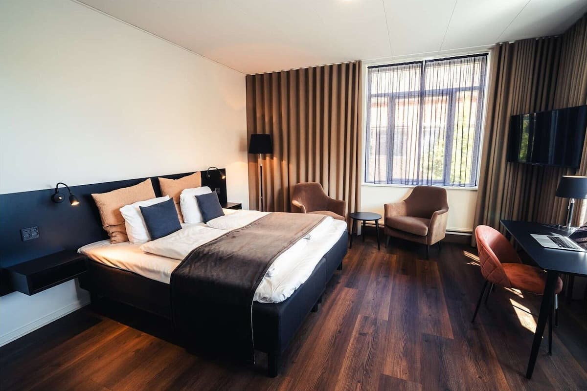 A hotel room with a large bed, two chairs, a desk with a laptop, a TV, and a window with curtains.