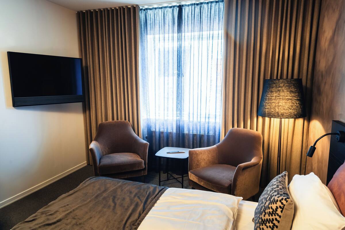 A hotel room with a bed, two armchairs, a small table, a TV mounted on the wall, and a lamp.