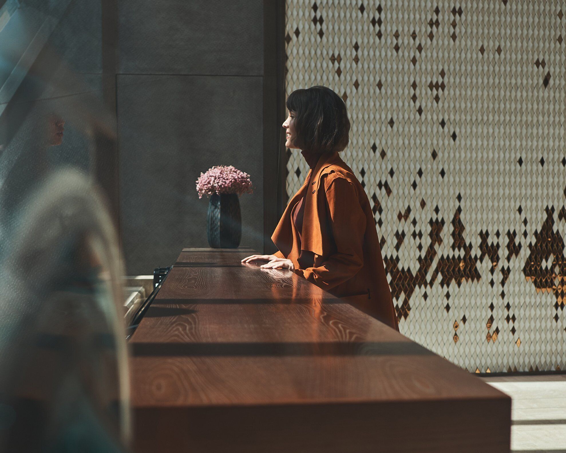 Una persona sentada a una mesa de madera con un jarrón de flores rosadas, frente a una pared con diseño.