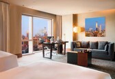 A hotel room with a large window offering a city view, a sofa, a desk with a lamp, and a painting on the wall.