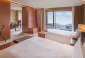 A hotel room with two beds, a desk, a chair, and a view of the city.