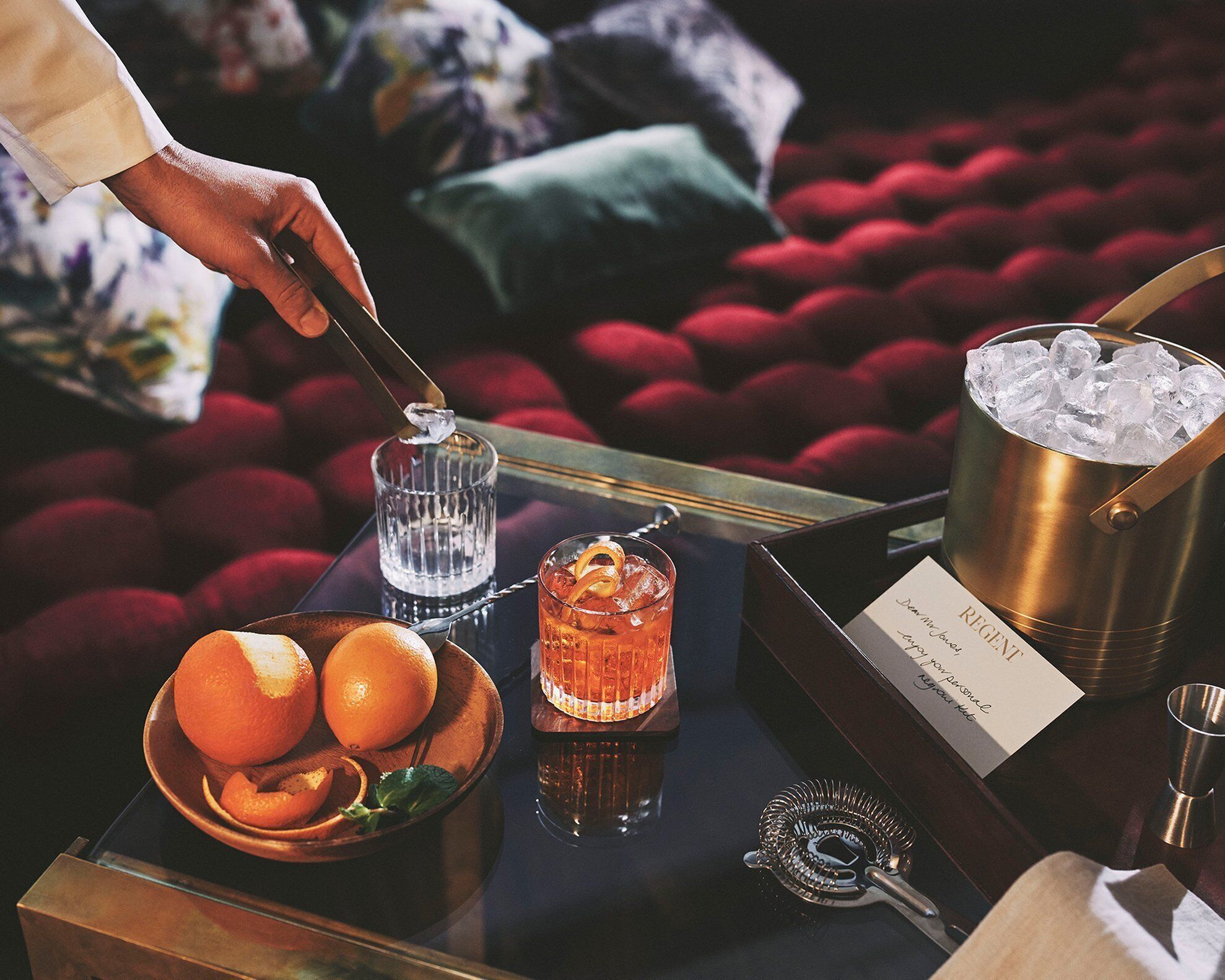 Una zona de bar con una bandeja que contiene un vaso de líquido ámbar, un cuenco de naranjas y una cuchara de metal para hielo.