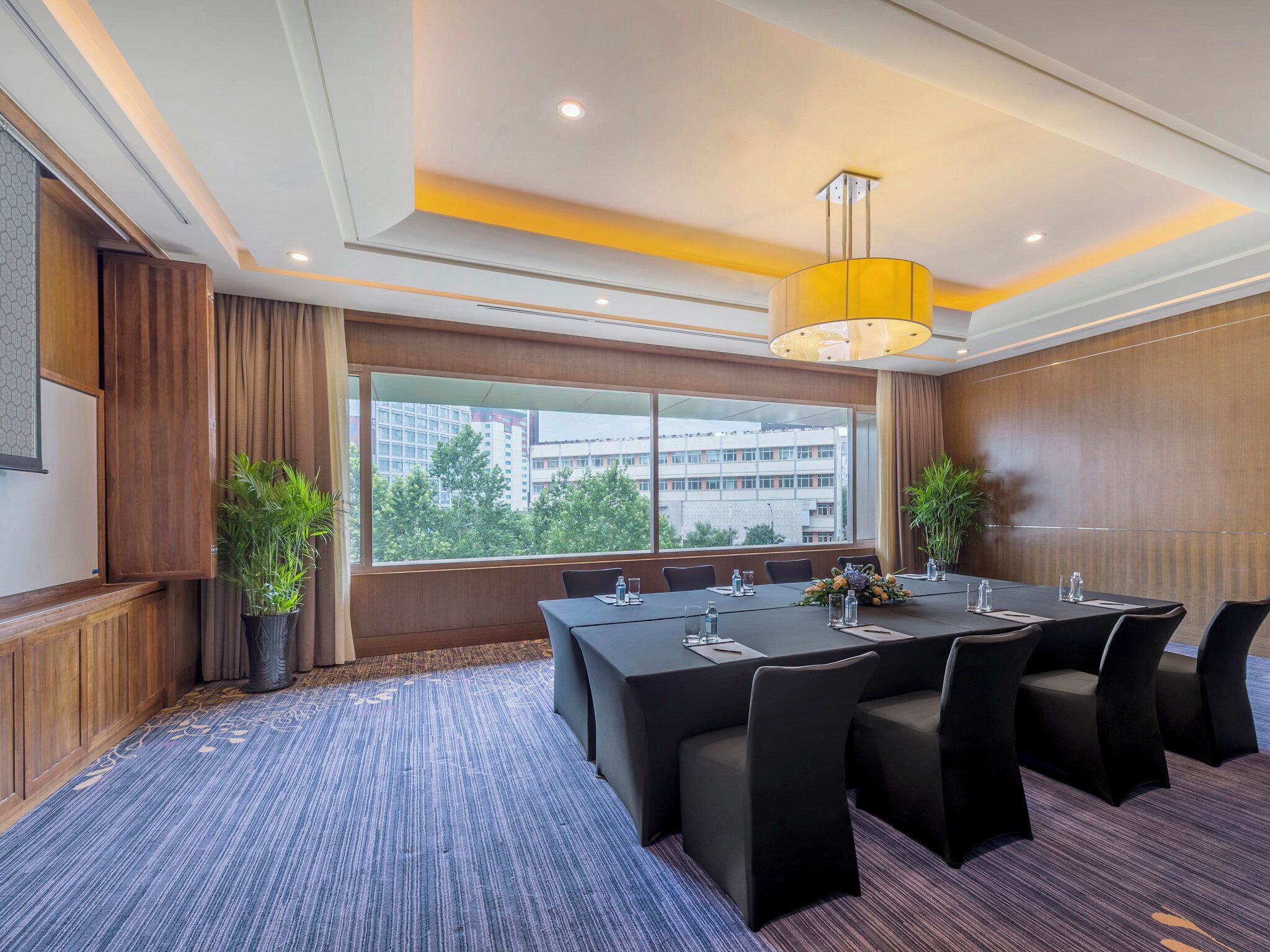 A conference room with a large table, chairs, a potted plant, and a view of buildings through the window.