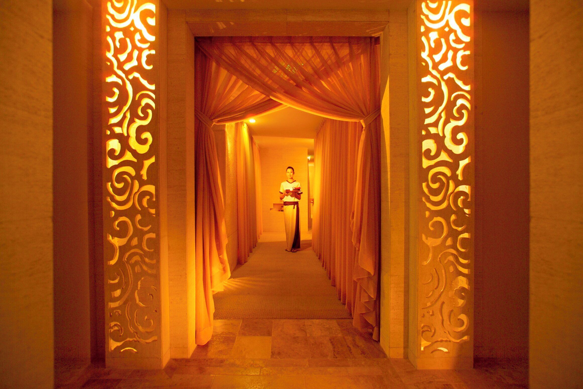 Un pasillo con columnas ornamentales y telas drapeadas, que conduce a una habitación donde hay una persona de pie.