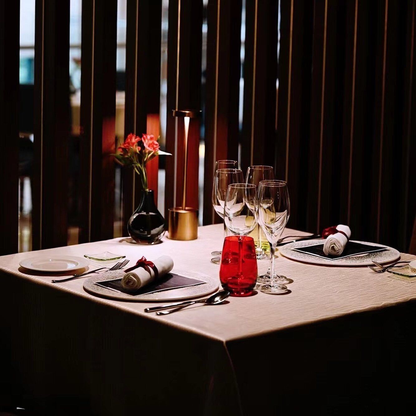 Una mesa de comedor dispuesta con platos, cubiertos y vasos, con un florero y una vela al fondo.