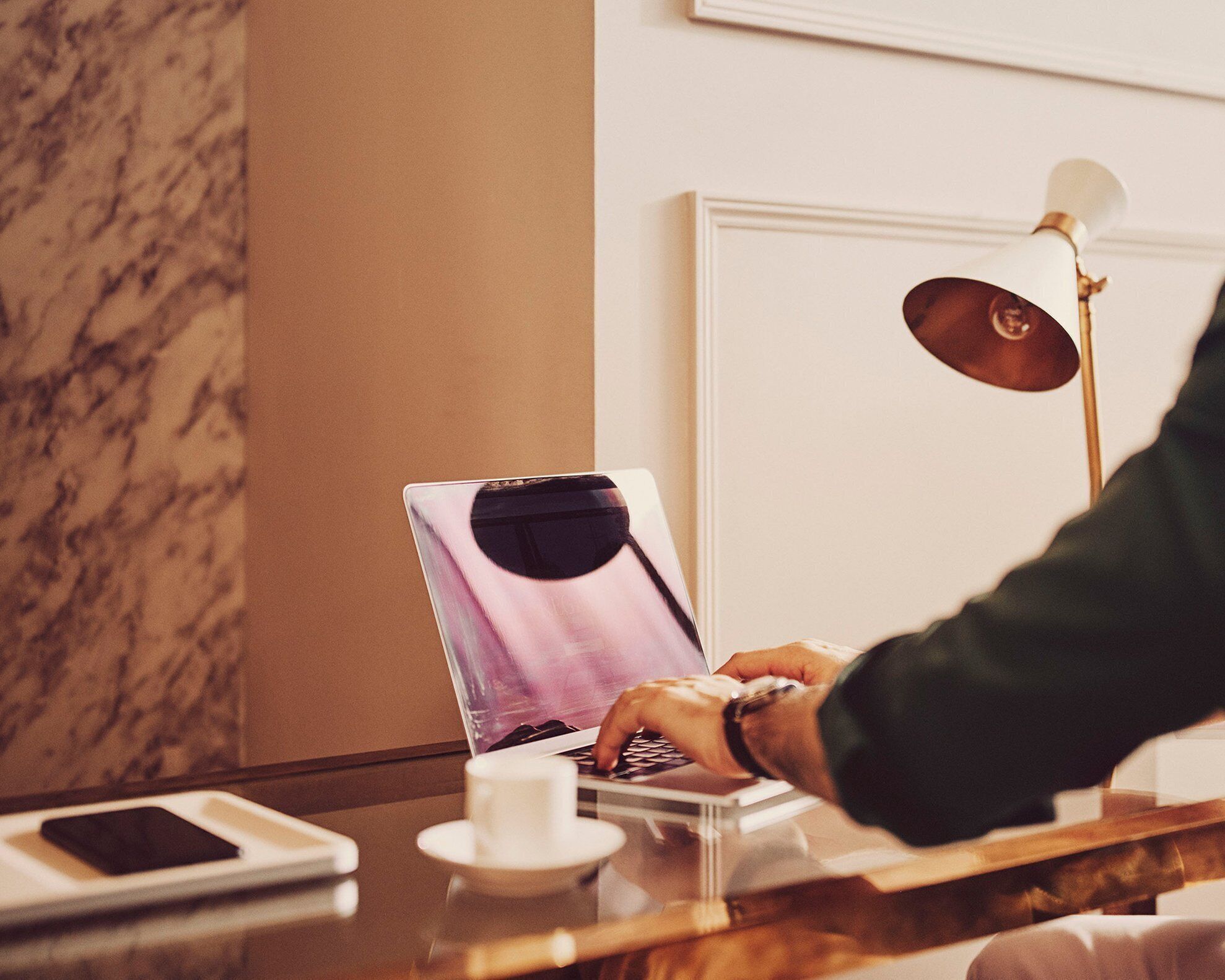 A laptop on a table with a smartphone and a cup, a lamp, and a marble wall.