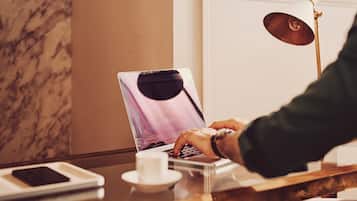 A laptop on a table with a smartphone and a cup, a lamp, and a marble wall.