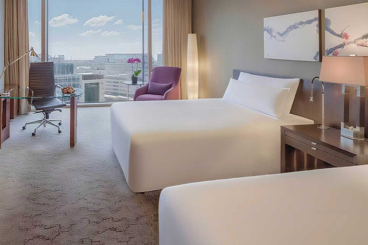 A hotel room with two beds, a desk with a chair, a lamp, and a view of the city.