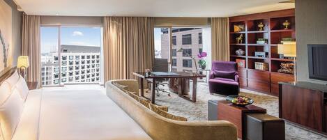 A hotel room with a large bed, a sofa, a desk, a chair, a bookshelf, and a television.