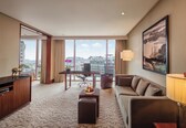 A hotel room with a large window, a sofa, a desk, and a dining table.