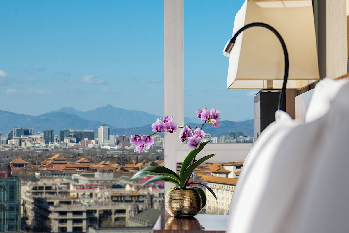 A cityscape view from a window with a vase of purple orchids on a table.
