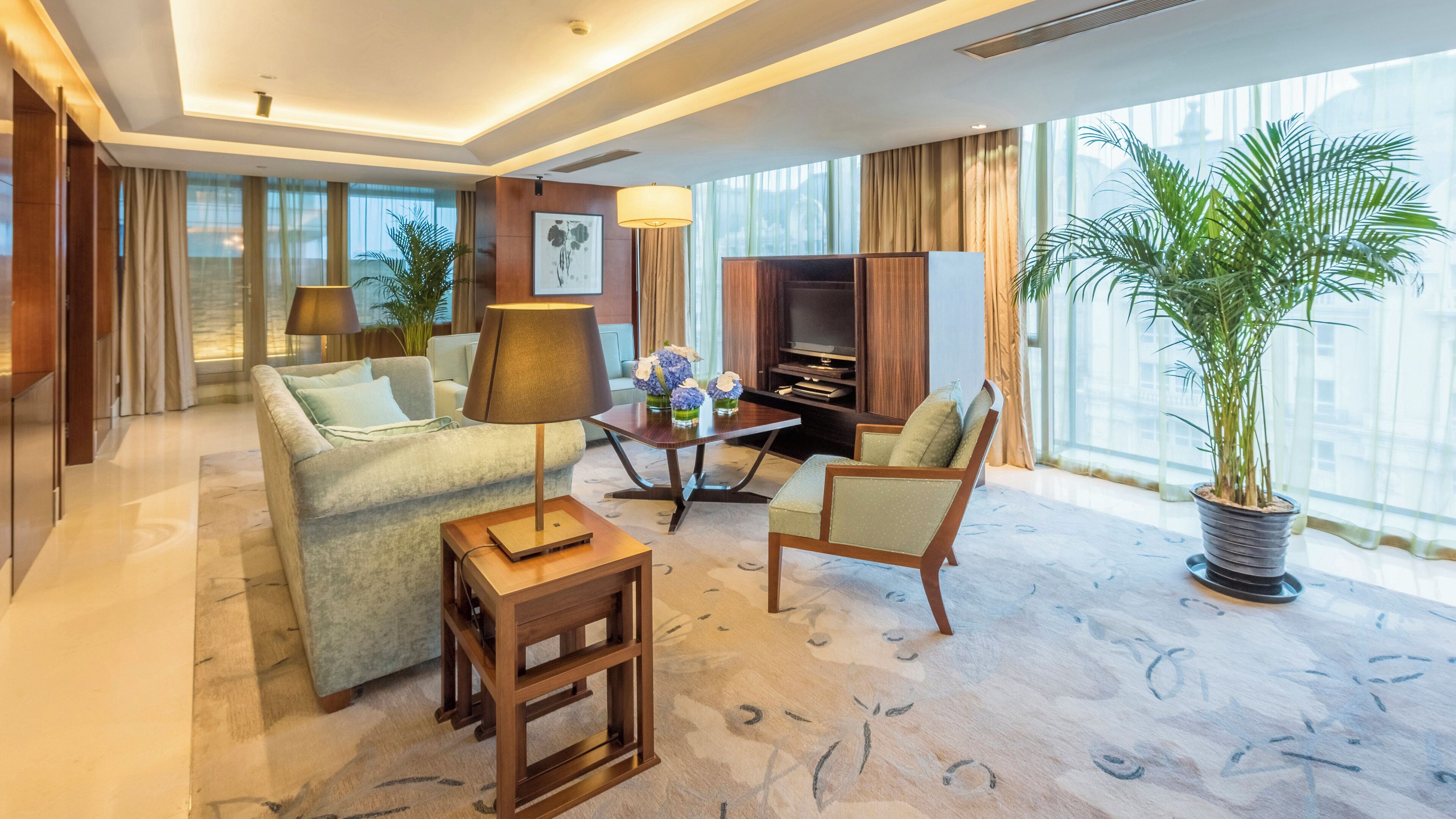 A hotel room with a sofa, armchair, TV, and a large potted plant.