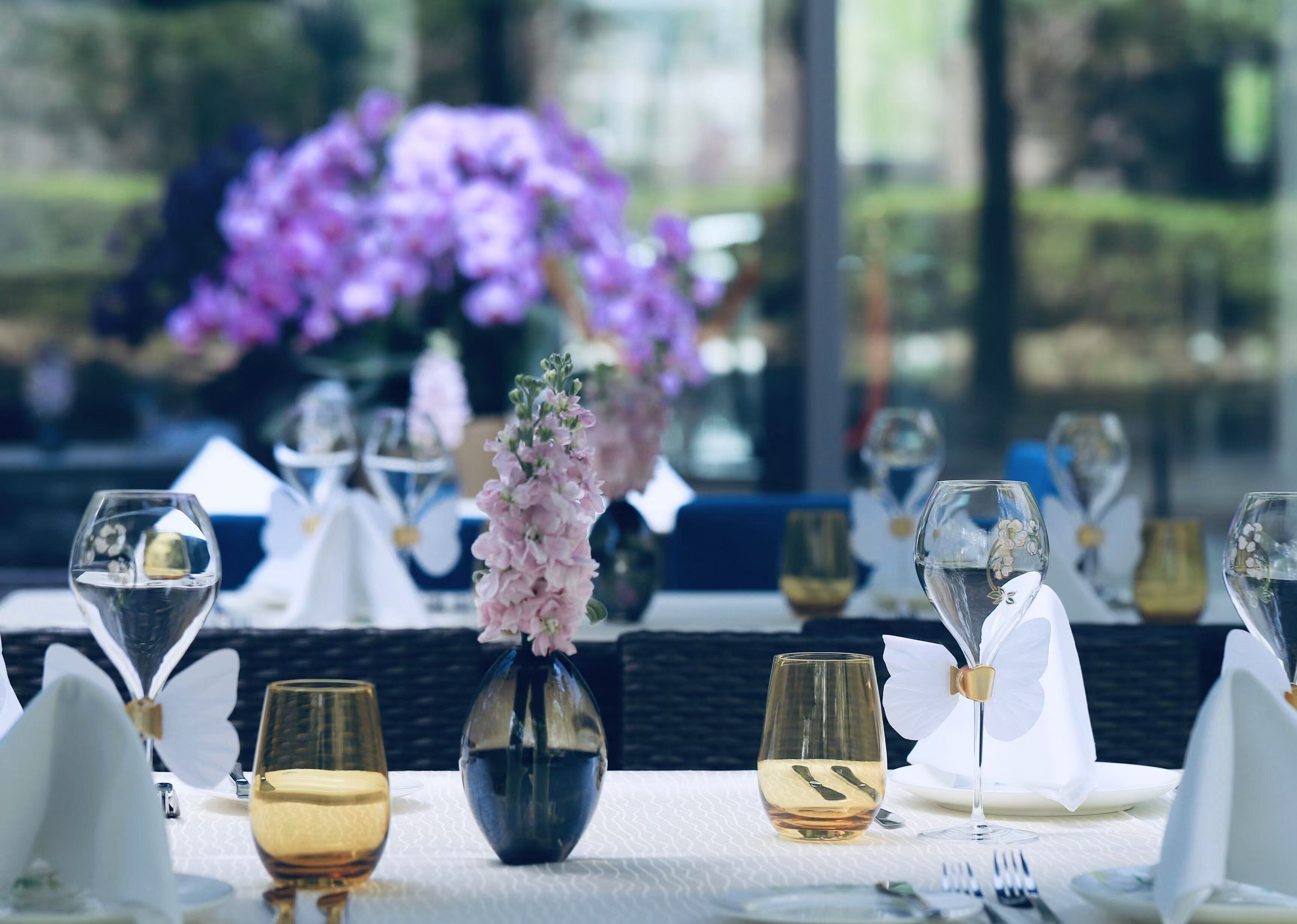 Una mesa de comedor preparada con copas de vino, un florero con flores y vistas a flores púrpuras a través de una ventana.