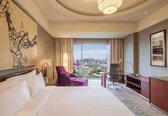 A hotel room with a large bed, a desk, a chair, a lamp, and a view of the city.