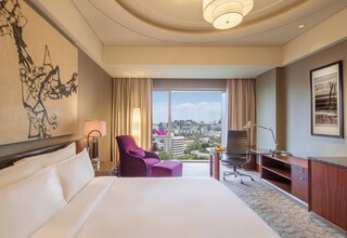 A hotel room with a large bed, a desk, a chair, a lamp, and a view of the city.