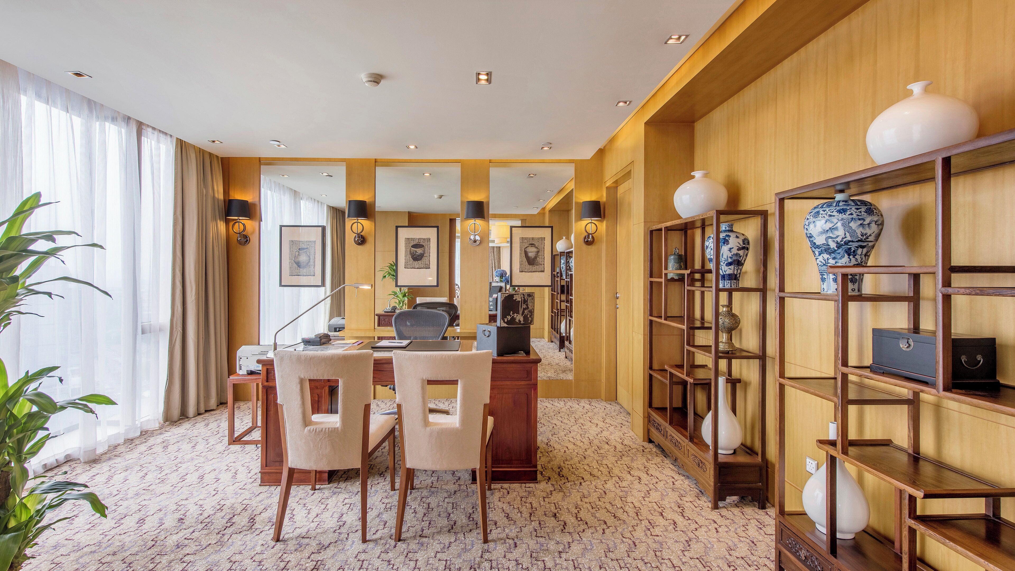 Habitación de hotel con escritorio de madera, sillas, una estantería y un ventanal con cortinas.