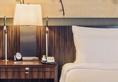 A hotel room with a wooden nightstand, a lamp, a clock, a telephone, and a bed with white bedding.