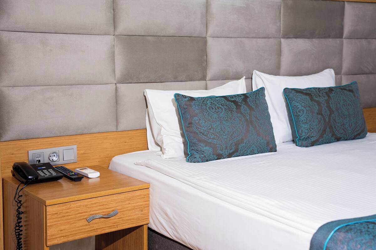 A hotel room with a bed, a bedside table with a phone and remote, and a wall socket.