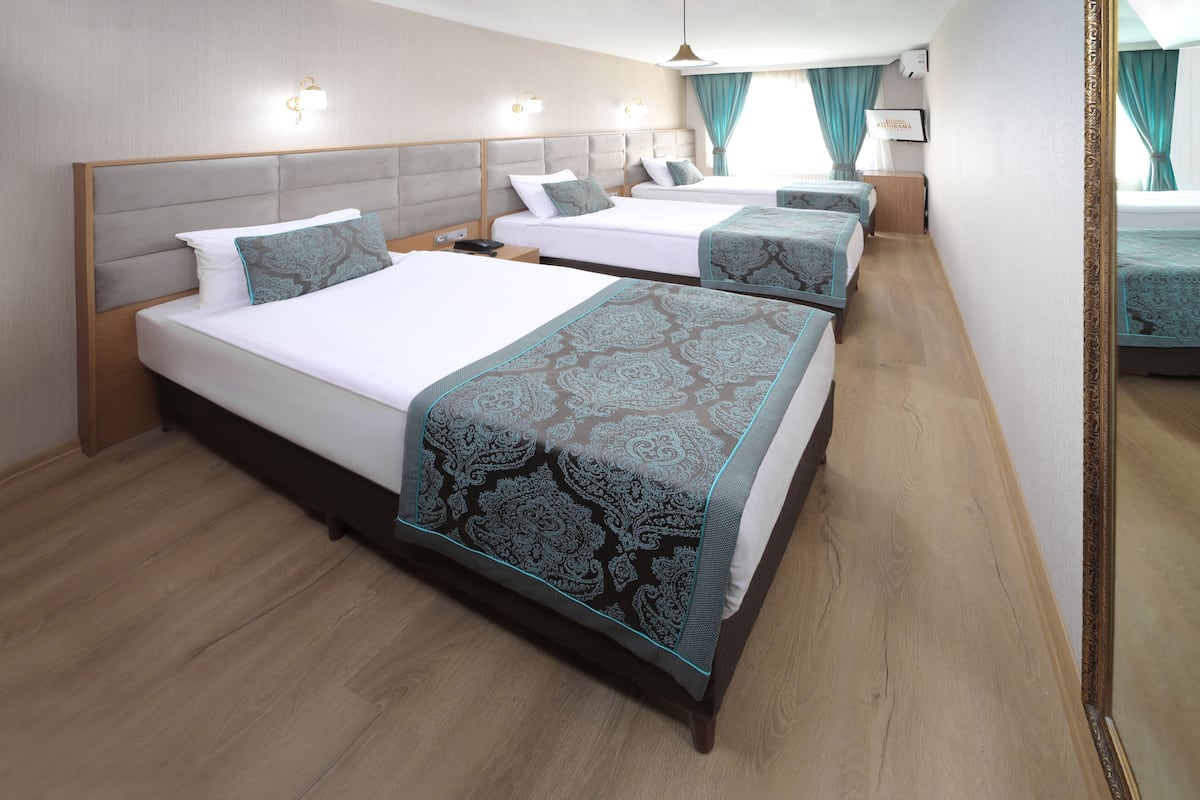 A hotel room with three beds, each with a patterned bedspread and matching pillows. The room has a wooden floor, a large mirror, and a ceiling light fixture.