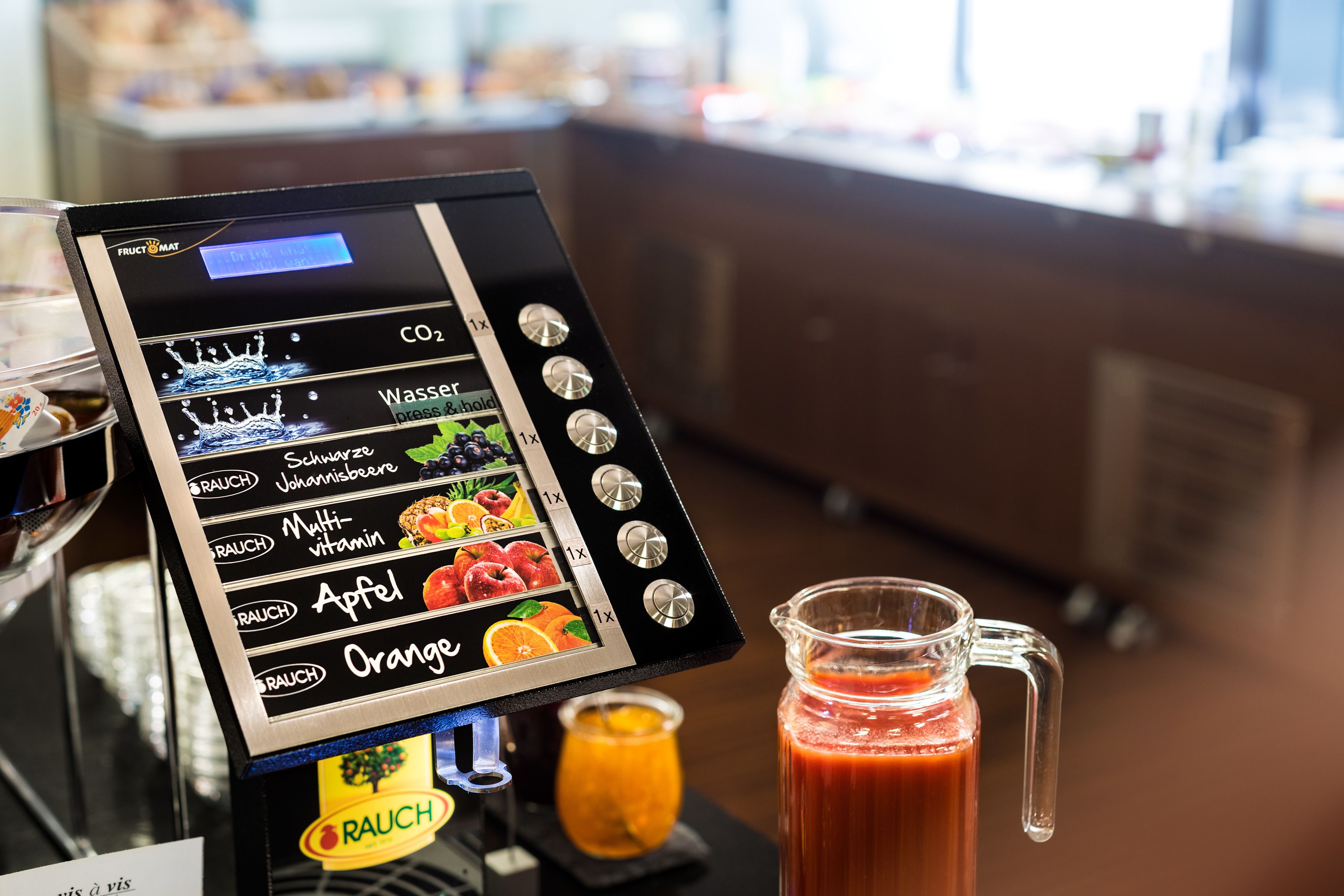 A coffee machine with a touchscreen interface displaying options for CO2, water, Schwarze Johannisbeeren, Multi-vitamin, Apfel, and Orange.