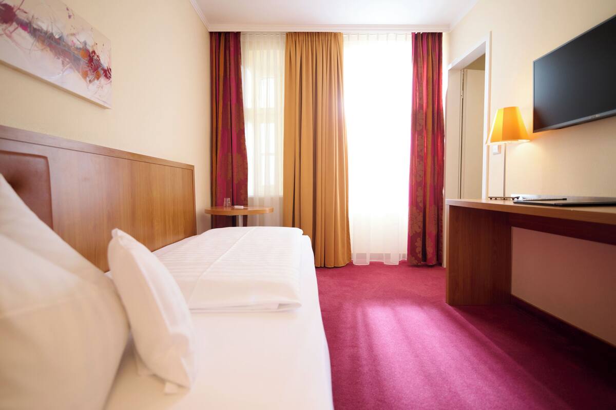 A hotel room with a bed, a desk, a TV, a lamp, and a window with curtains.