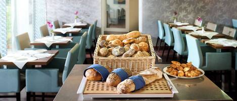 A restaurant interior with tables, chairs, a basket of bread, and a plate of pastries.