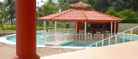 Un área de piscina con un pabellón, mobiliario exterior blanco y palmeras al fondo.