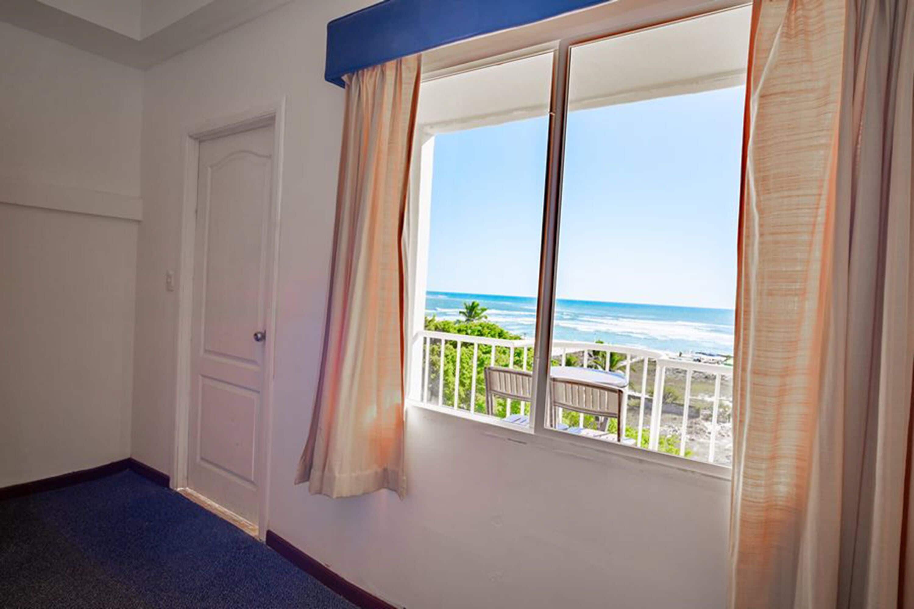 A room with a large window overlooking a beach, a door, and a blue carpet.
