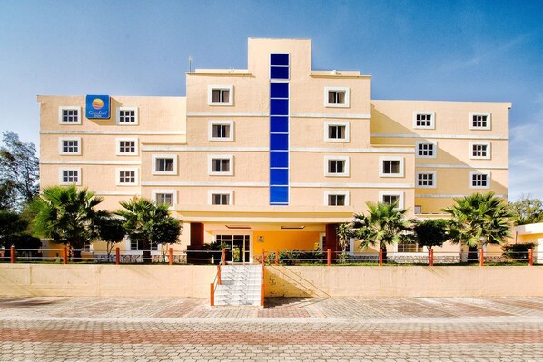 Un hôtel à plusieurs étages, doté d’un élément vertical bleu central, d’un toit plat et d’une zone d’entrée pavée agrémentée de palmiers.