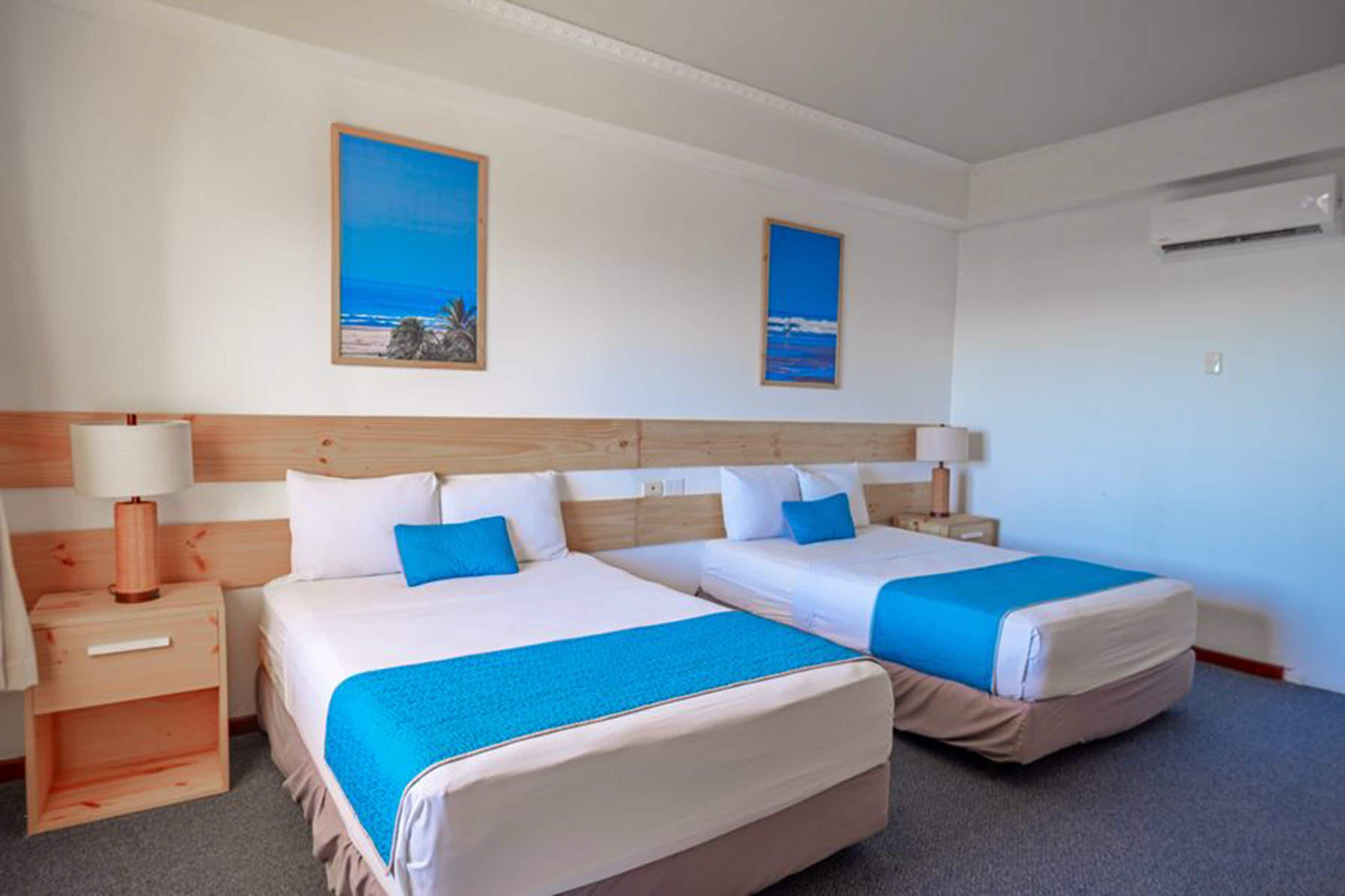 A hotel room with two beds, a wooden headboard, a nightstand with a lamp, and two framed pictures on the wall.