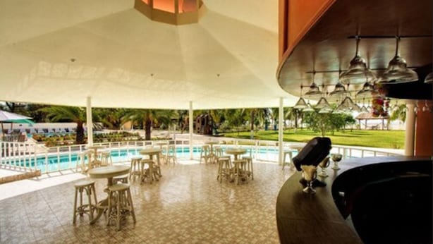 Un área junto a la piscina con varias mesas y sillas, una zona de bar y vistas a una piscina y follaje.