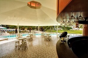 A poolside area with multiple tables and chairs, a bar area, and a view of a pool and greenery.