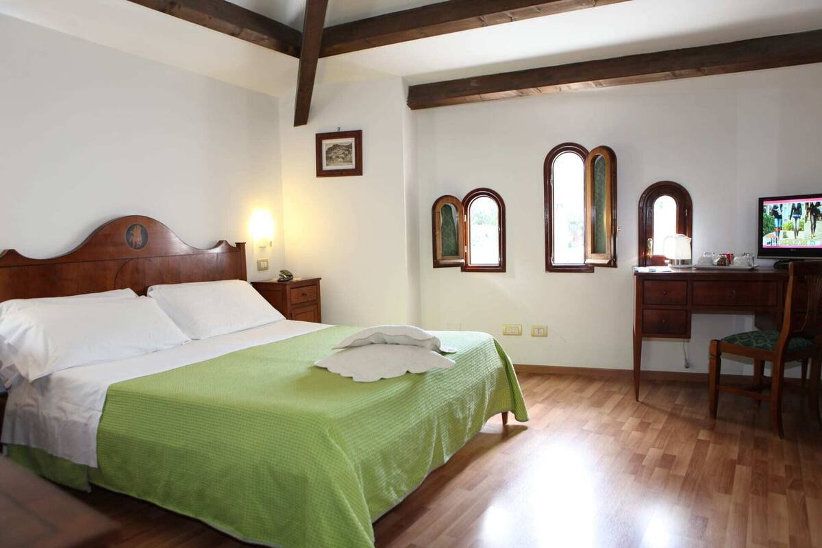 Une chambre à coucher avec un lit en bois, un bureau avec une chaise, une télévision et deux fenêtres en arc de cercle.