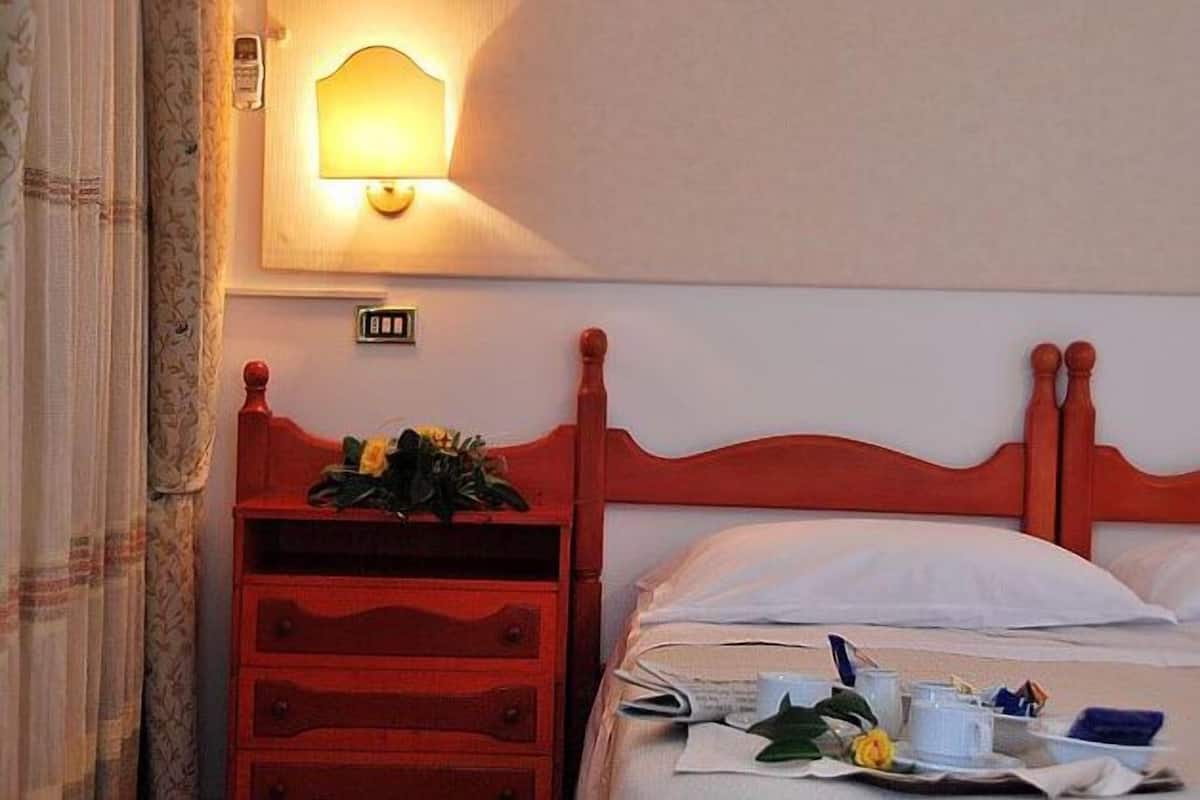A hotel room with a wooden headboard, a bedside table with a lamp, and a bed with a white pillow and a floral arrangement.