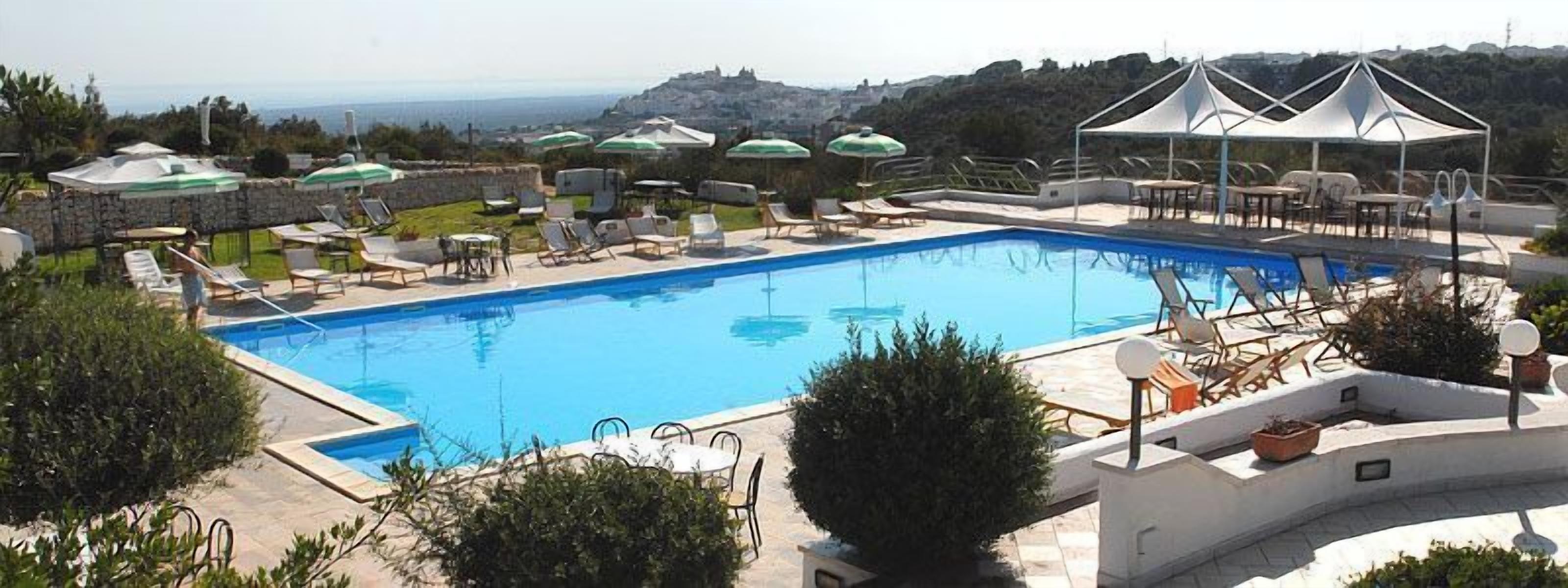 A pool with lounge chairs and umbrellas, surrounded by stone walls and greenery, with a view of hills in the background.