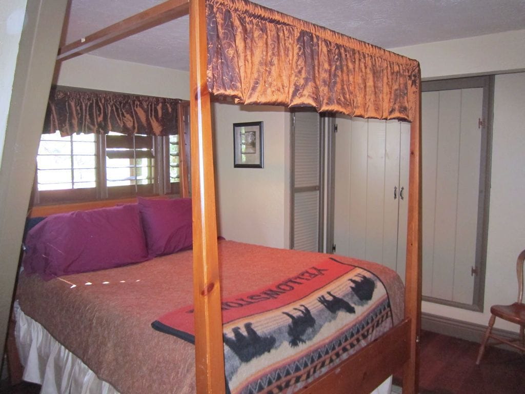 A four-poster bed with a patterned bedspread and pillows, a wooden headboard, and a window with curtains.